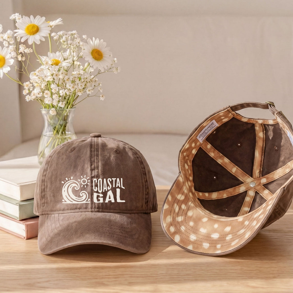 Two baseball caps displayed on a wooden table in a styled indoor setting. One cap is front-facing and appears dusty brown with a white 'COASTAL GAL' logo on the front.