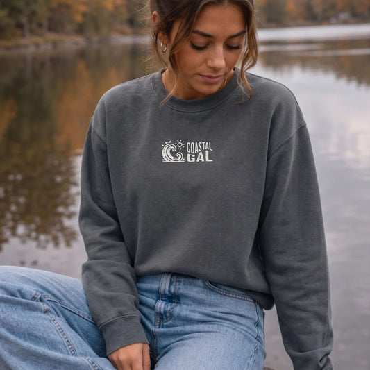 A woman sitting by a lake wearing a dark gray crewneck sweatshirt with a white 'COASTAL GAL' wave graphic on the chest, paired with light blue jeans.