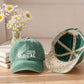 A green baseball cap with white 'COASTAL GAL' text and a daisy-and-beige floral pattern on the brim, shown alongside the inside view of the cap. The cap is styled on a wooden table next to books and a vase of daisies in front of a beige sofa.