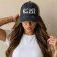 A woman wearing a dark washed trucker hat with a white 'COASTAL GAL' wave graphic on the front, paired with a white t-shirt against a plain beige background.