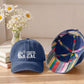 Two navy blue baseball caps displayed on a wooden table. The left cap is solid navy with a white 'COASTAL GAL' wave logo on the front, resting on stacked books next to a vase of white daisies.