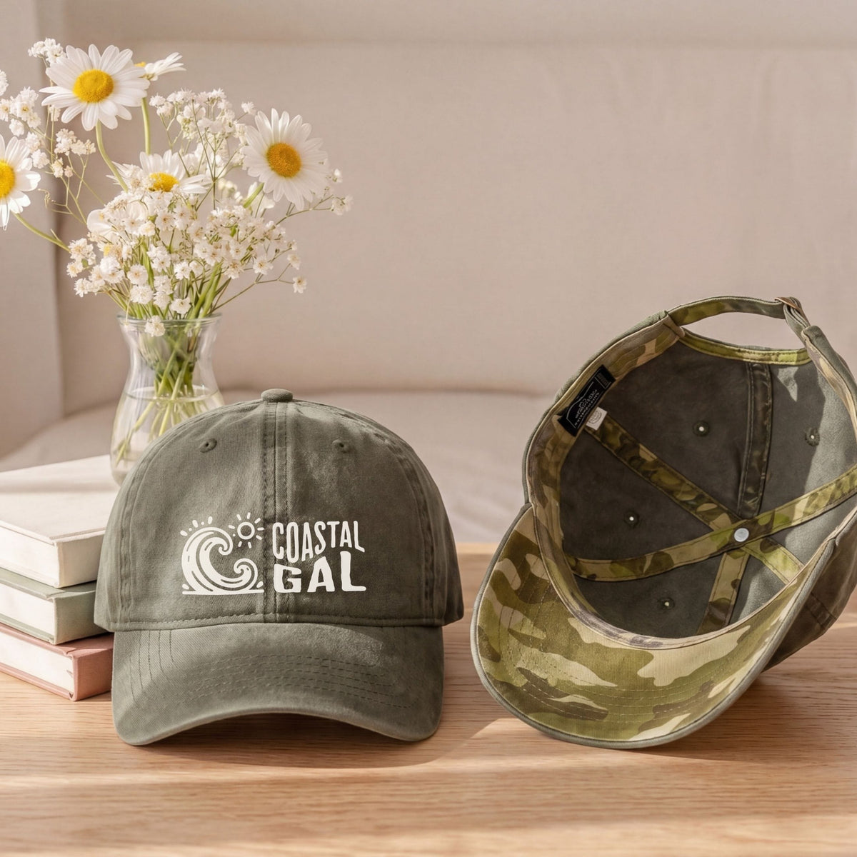 Two baseball caps displayed on a wooden table with a vase of white daisies and stacked books in the background. The cap on the left is olive green with a white 'COASTAL GAL' wave logo on the front.