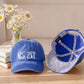 A blue baseball cap shown from the front and side on a wooden table, with a vase of white daisies and stacked books in the background. The front of the cap has a white 'COASTAL GAL' wave logo, and the inside of the cap features a glittery blue and silver pattern.