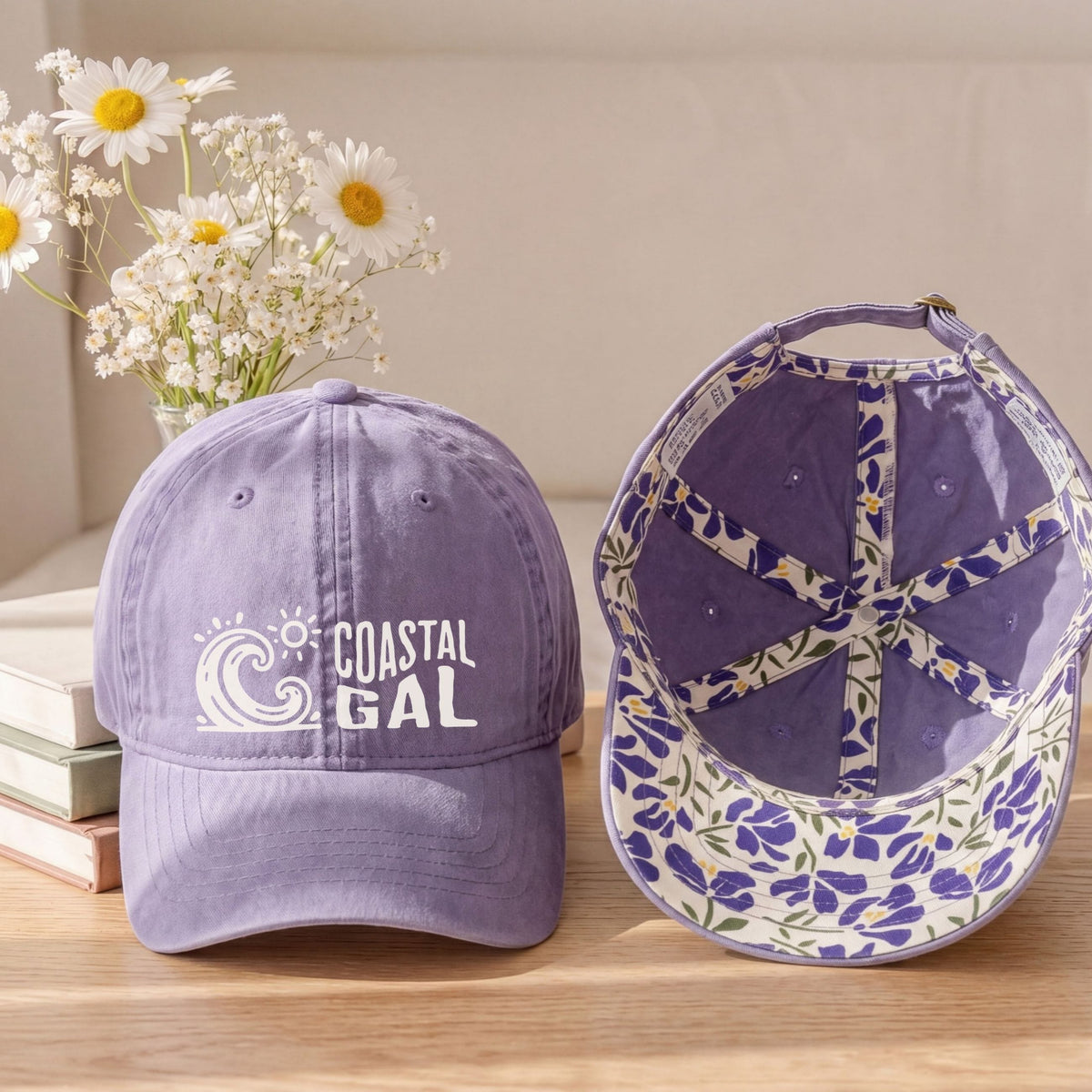 A lavender purple baseball cap shown from the front and inside, placed on a wooden surface with stacked books and a vase of white daisies in the background. The front has a white 'COASTAL GAL' wave graphic, and the inside features a floral pattern in purple, white, and green.