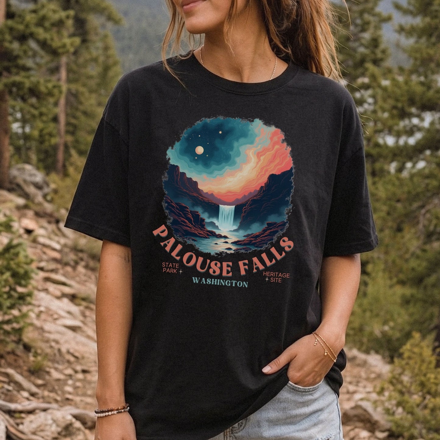 A woman wearing a black graphic t-shirt in an outdoor forest setting. The shirt features a colorful circular print of a waterfall and mountains under a starry sky, with the text 'PALOUSE FALLS WASHINGTON' printed below.