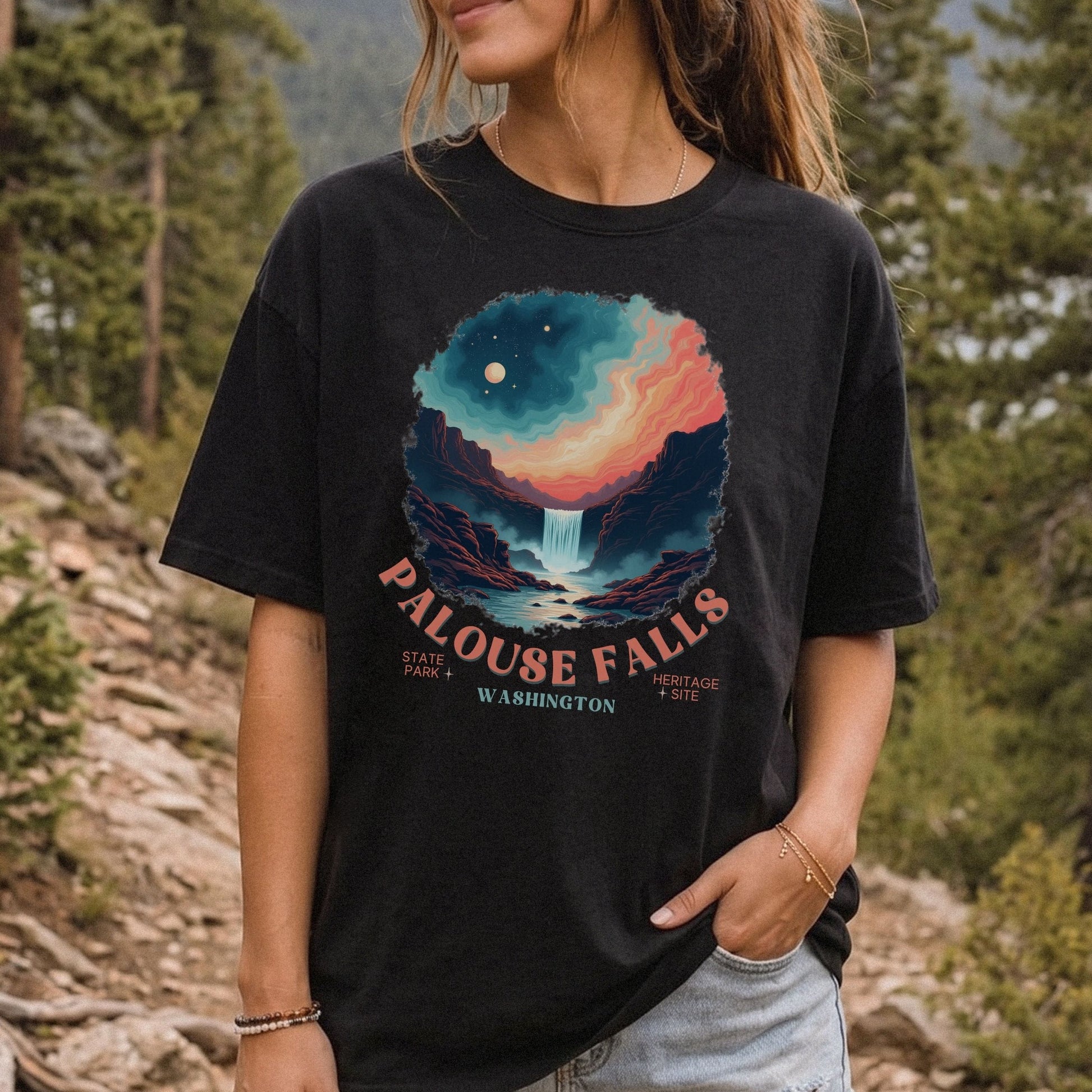 A woman wearing a black graphic t-shirt in an outdoor forest setting. The shirt features a colorful circular print of a waterfall and mountains under a starry sky, with the text 'PALOUSE FALLS WASHINGTON' printed below.