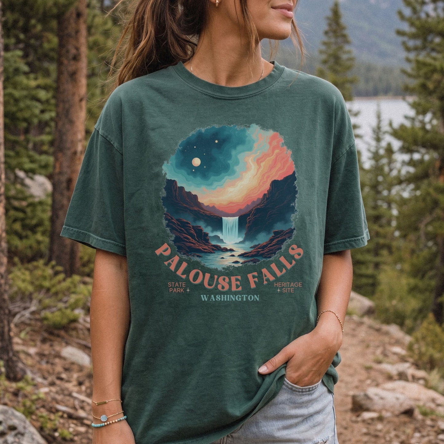 A woman wearing a dark green graphic t-shirt with a scenic Palouse Falls print, standing outdoors in a forest setting.