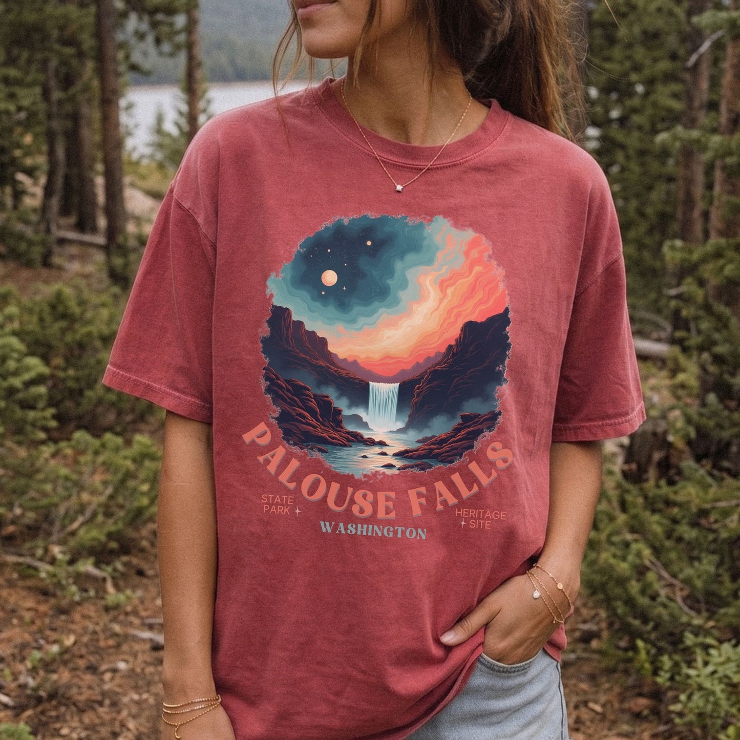 A woman wearing a faded dusty rose graphic t-shirt with a colorful Palouse Falls landscape print, standing in a forest setting.