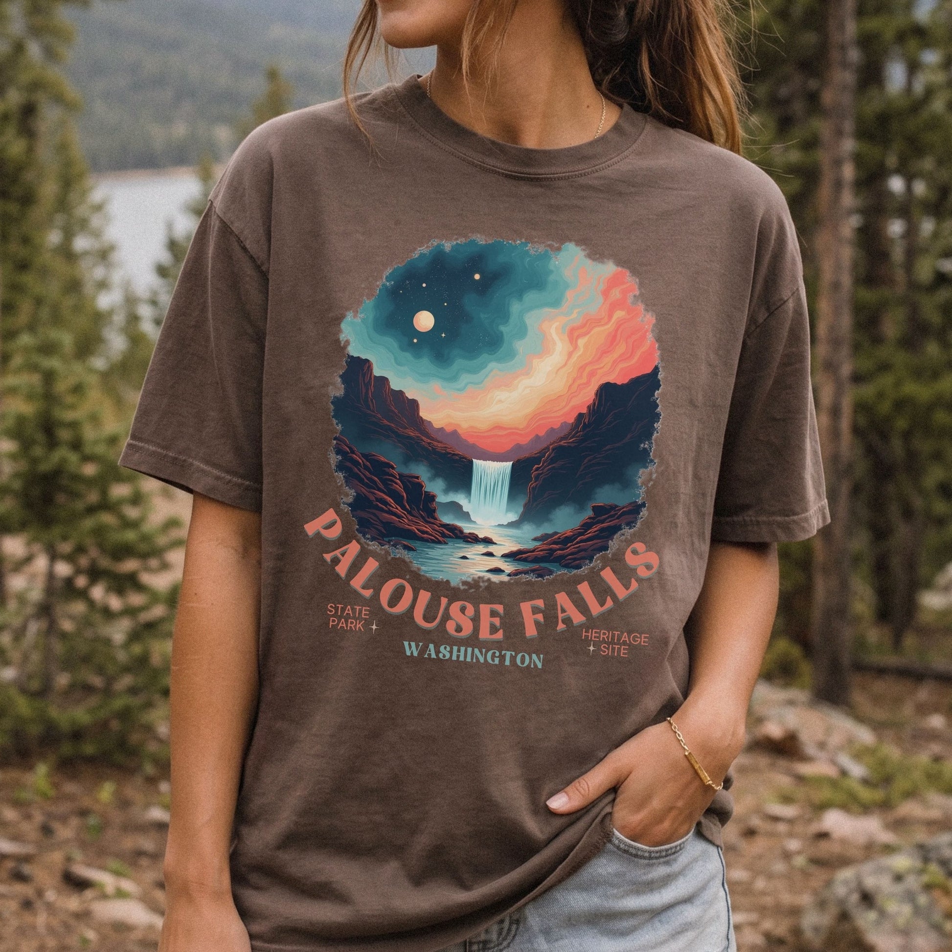 A woman wearing a brown graphic t-shirt in an outdoor forest setting. The t-shirt features a colorful circular print of a waterfall in Palouse Falls, Washington, with the text 'PALOUSE FALLS' and 'WASHINGTON'.