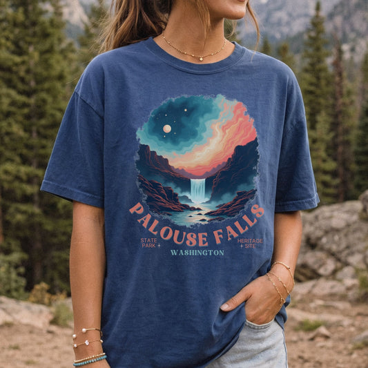 A woman wearing a navy blue graphic t-shirt with a colorful Palouse Falls landscape print, standing outdoors in a forest setting.