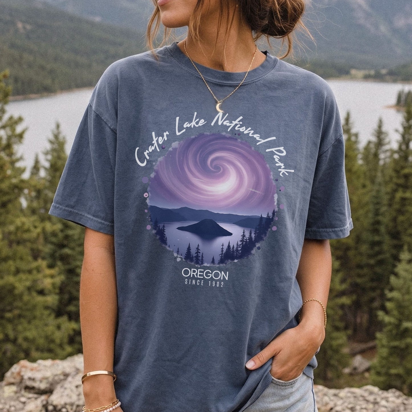 A woman wearing a Comfort Colors Denim t-shirt with a vintage-style circular graphic of Crater Lake at night under a purple spiral galaxy, framed by white cursive text that reads Crater Lake National Park Oregon.