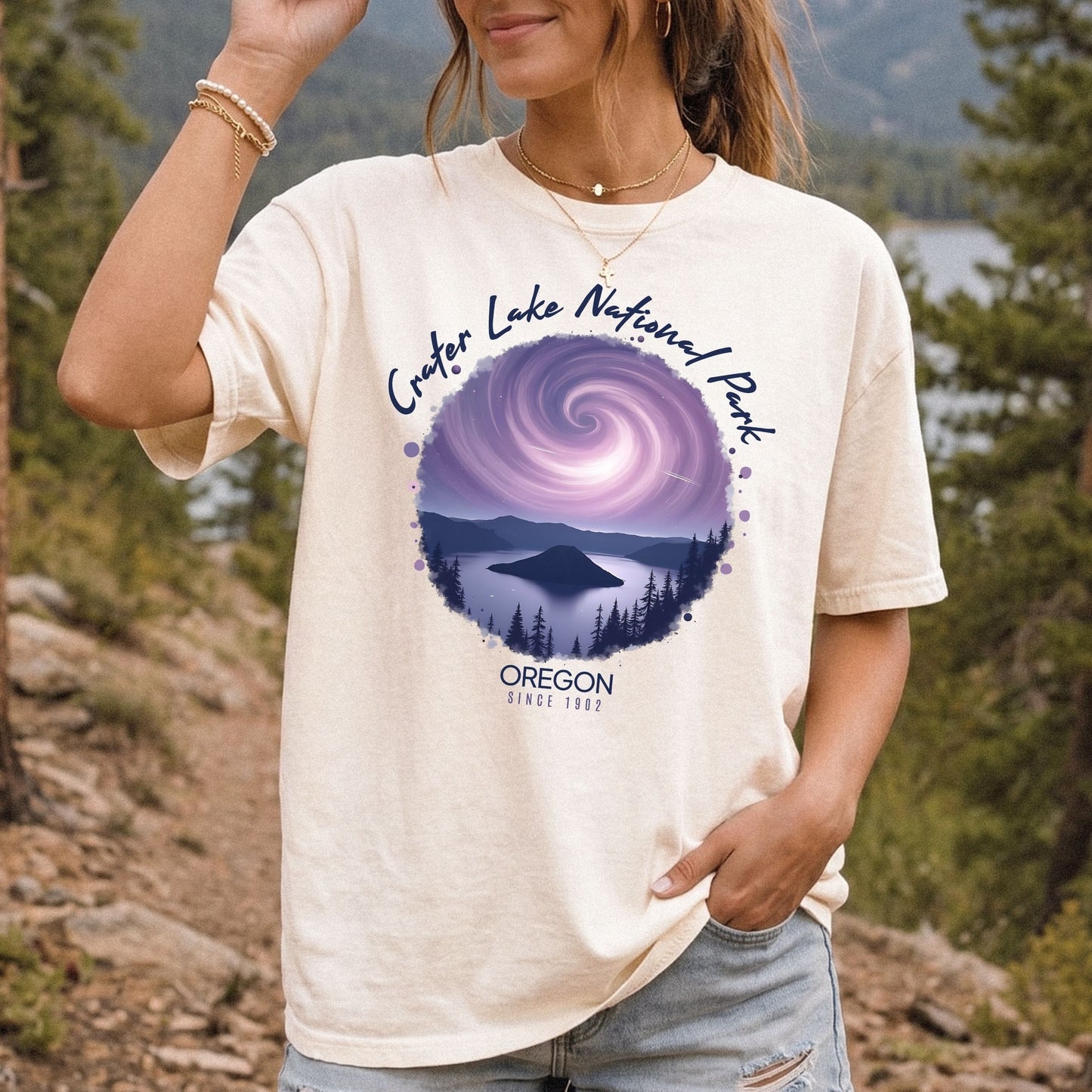 A woman wearing a Comfort Colors Ivory t-shirt with a vintage-style circular graphic of Crater Lake at night under a purple spiral galaxy, framed by white cursive text that reads Crater Lake National Park Oregon.