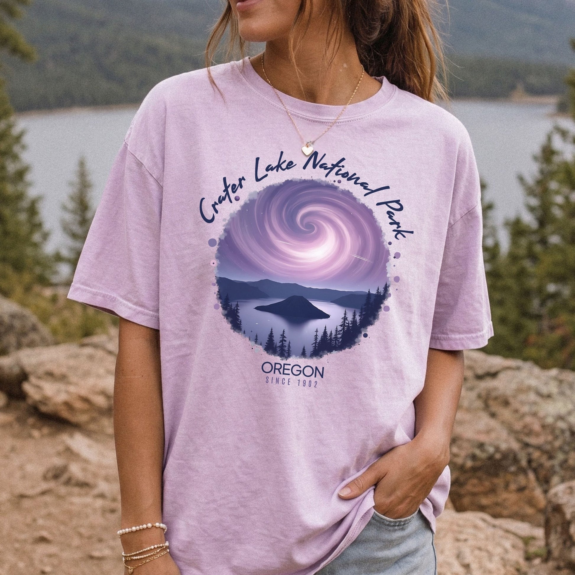 A woman wearing a Comfort Colors orchid t-shirt with a vintage-style circular graphic of Crater Lake at night under a purple spiral galaxy, framed by white cursive text that reads Crater Lake National Park Oregon.