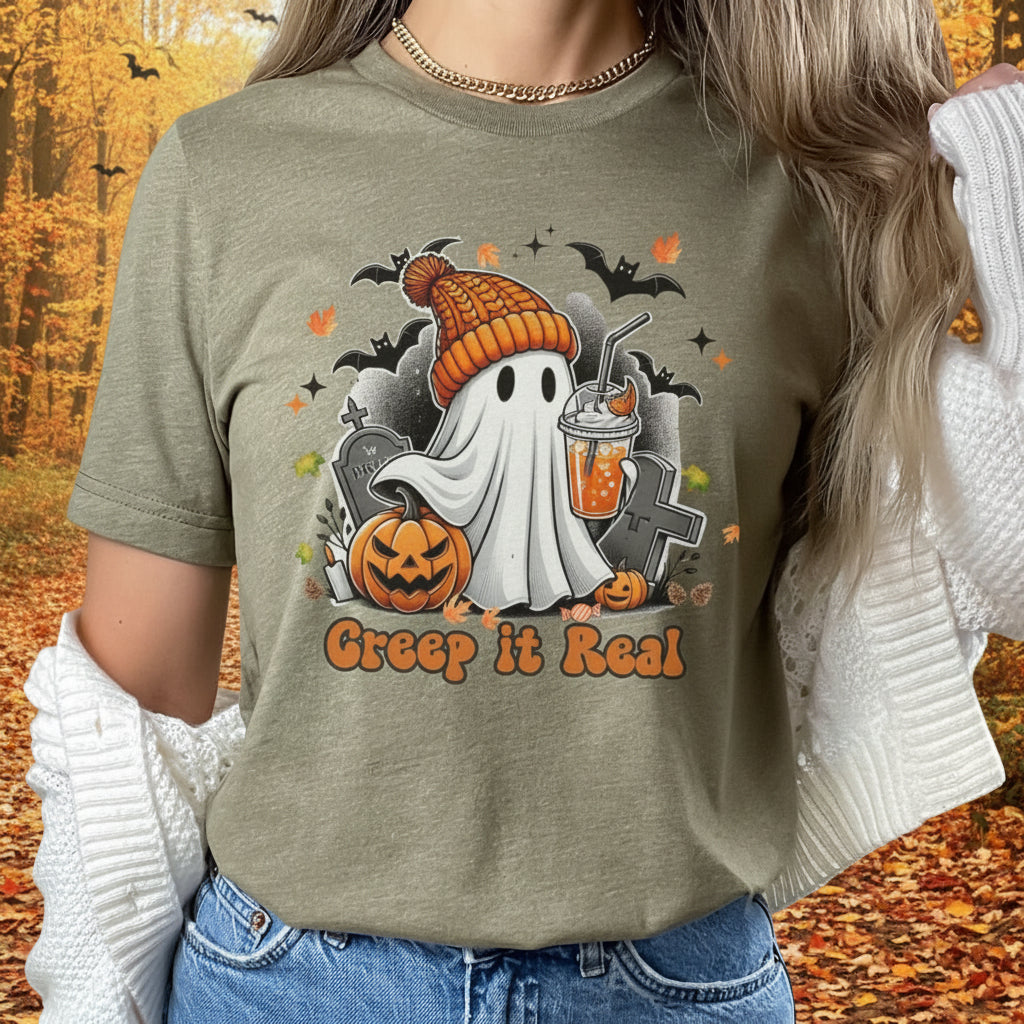 A woman wearing a muted olive green graphic t-shirt with a Halloween design featuring a ghost in an orange beanie, a jack-o'-lantern, a tombstone, and the text 'Creep it Real'. She is also wearing a white knit cardigan and blue jeans, set against a blurred autumn forest background.