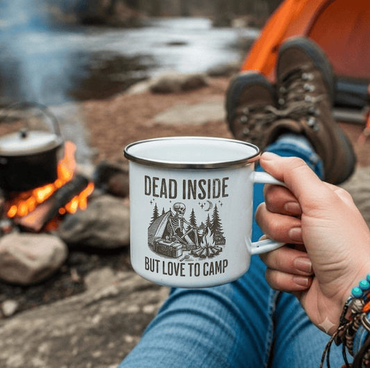 A person at a campfire by a river holds a white enamel mug with a brown camping skeleton graphic and the text 'DEAD INSIDE BUT LOVE TO CAMP'. The person's legs, hand, and hiking boots are visible, with a tent and flowing water in the background.