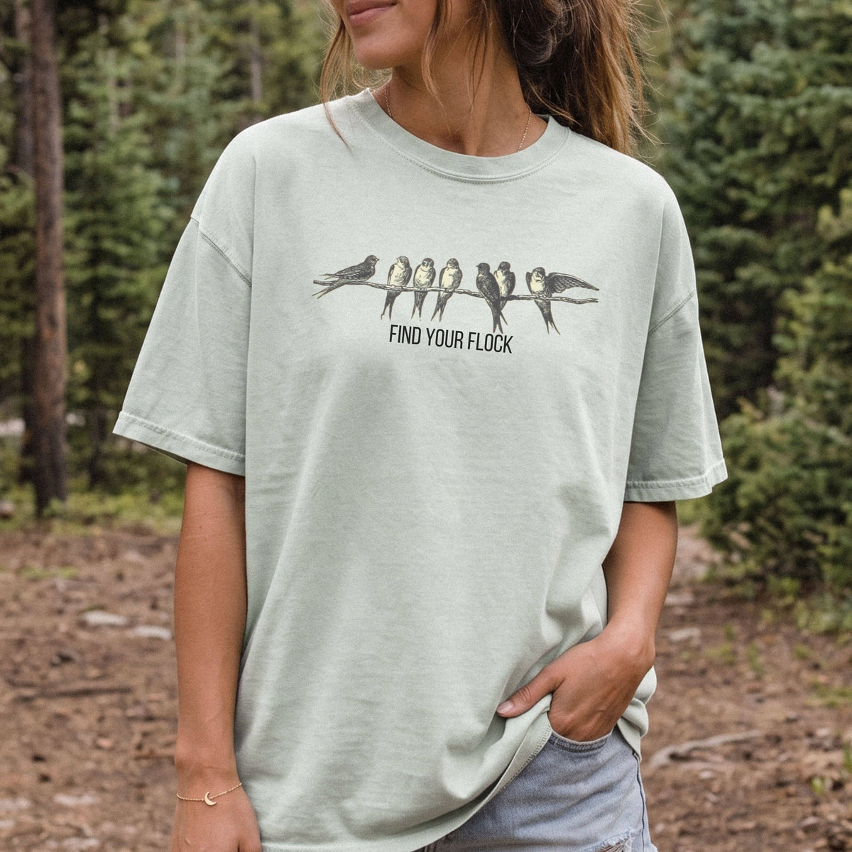 A woman wearing a light sage green graphic t-shirt with a black and white bird design and the text 'FIND YOUR FLOCK' on the front, set against a blurred forest background.