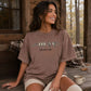 A woman sitting on a wooden porch wearing a brown graphic t-shirt with a bird design and the text 'FIND YOUR FLOCK'.