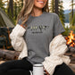 A woman sitting outdoors by a campfire wearing a heather gray graphic t-shirt with a bird design and the text 'FIND YOUR FLOCK'. She has a cream cable-knit sweater draped off her shoulders, black pants, and is holding a white mug.