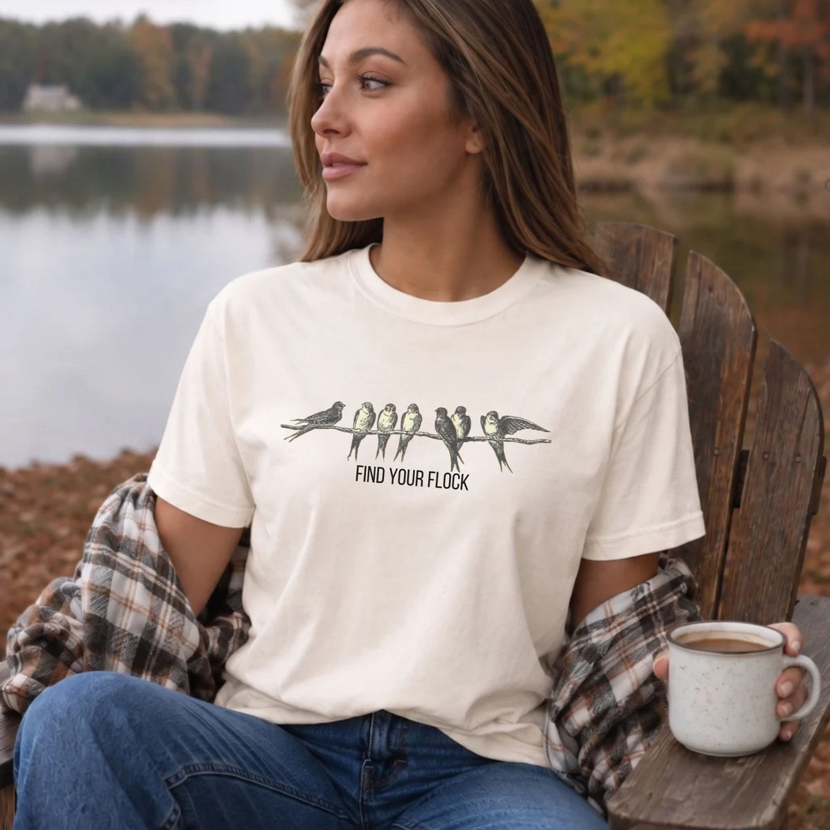 A woman sitting on a wooden chair by a lake wearing a cream graphic t-shirt with a bird print and the text 'FIND YOUR FLOCK'. She has a plaid shirt draped off her shoulders and is holding a mug.