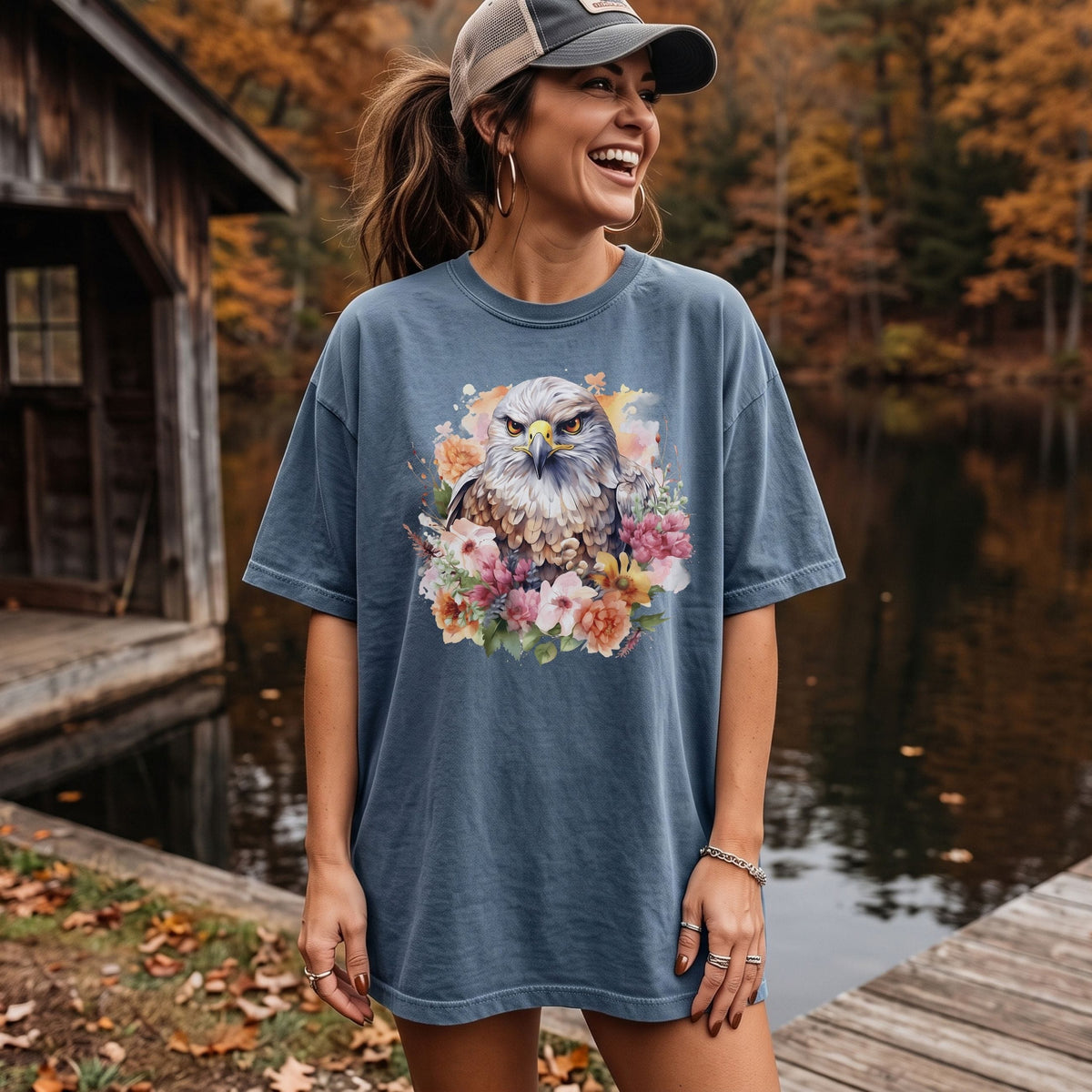 A woman wearing an oversized slate blue t-shirt with a colorful watercolor graphic of a hawk surrounded by flowers, standing on a wooden dock by a lake with autumn trees in the background.