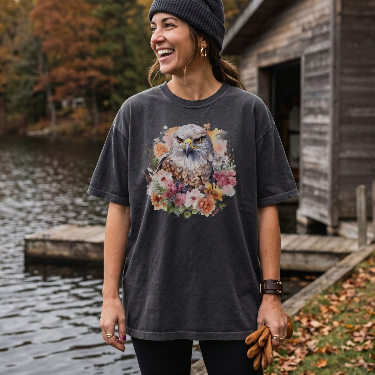 A woman wearing a dark charcoal gray graphic t-shirt with a detailed print of a hawk surrounded by colorful flowers, standing outdoors near a lake and a rustic wooden cabin.