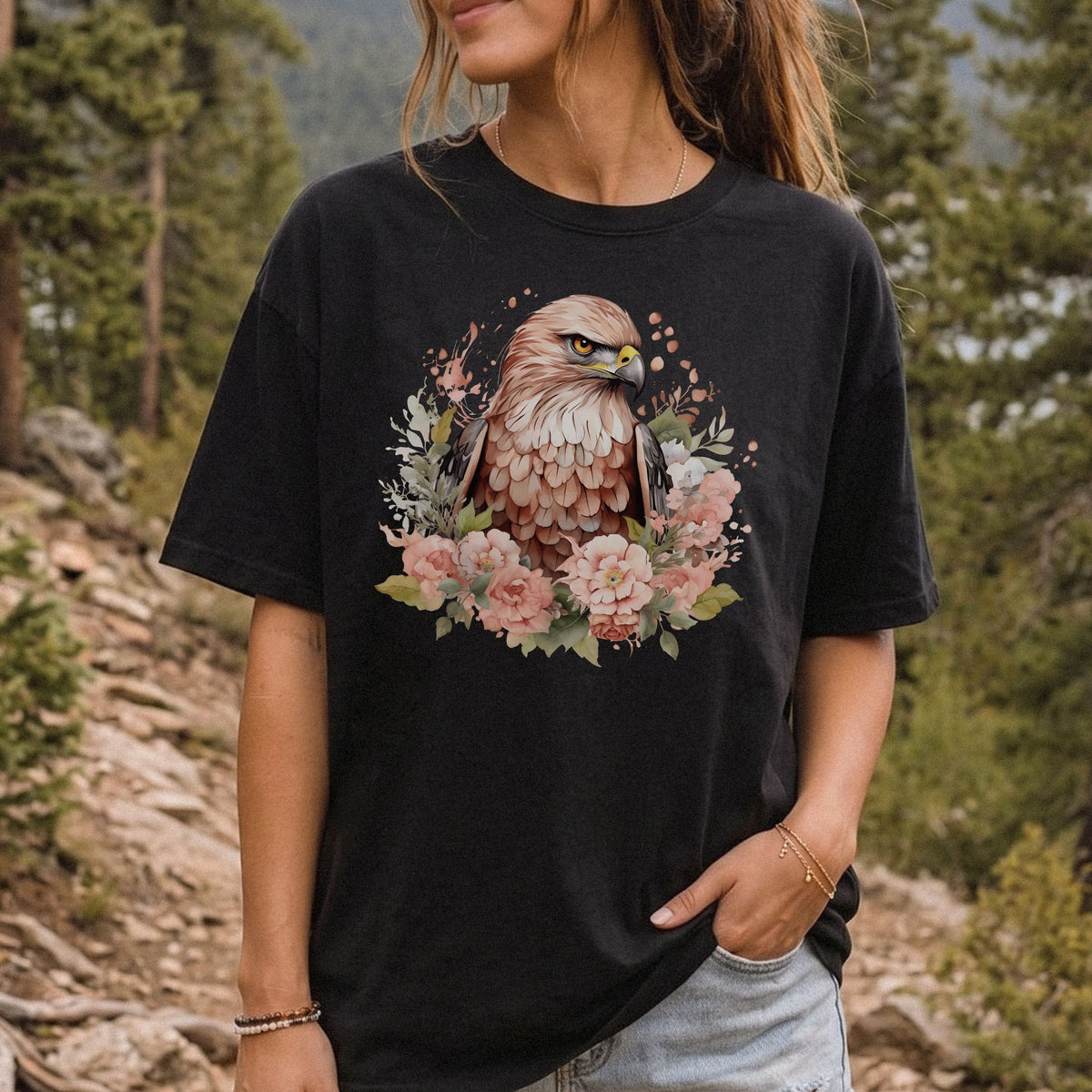 A woman wearing a black graphic t-shirt featuring a detailed illustration of a hawk surrounded by pink flowers, set against a blurred forest background.