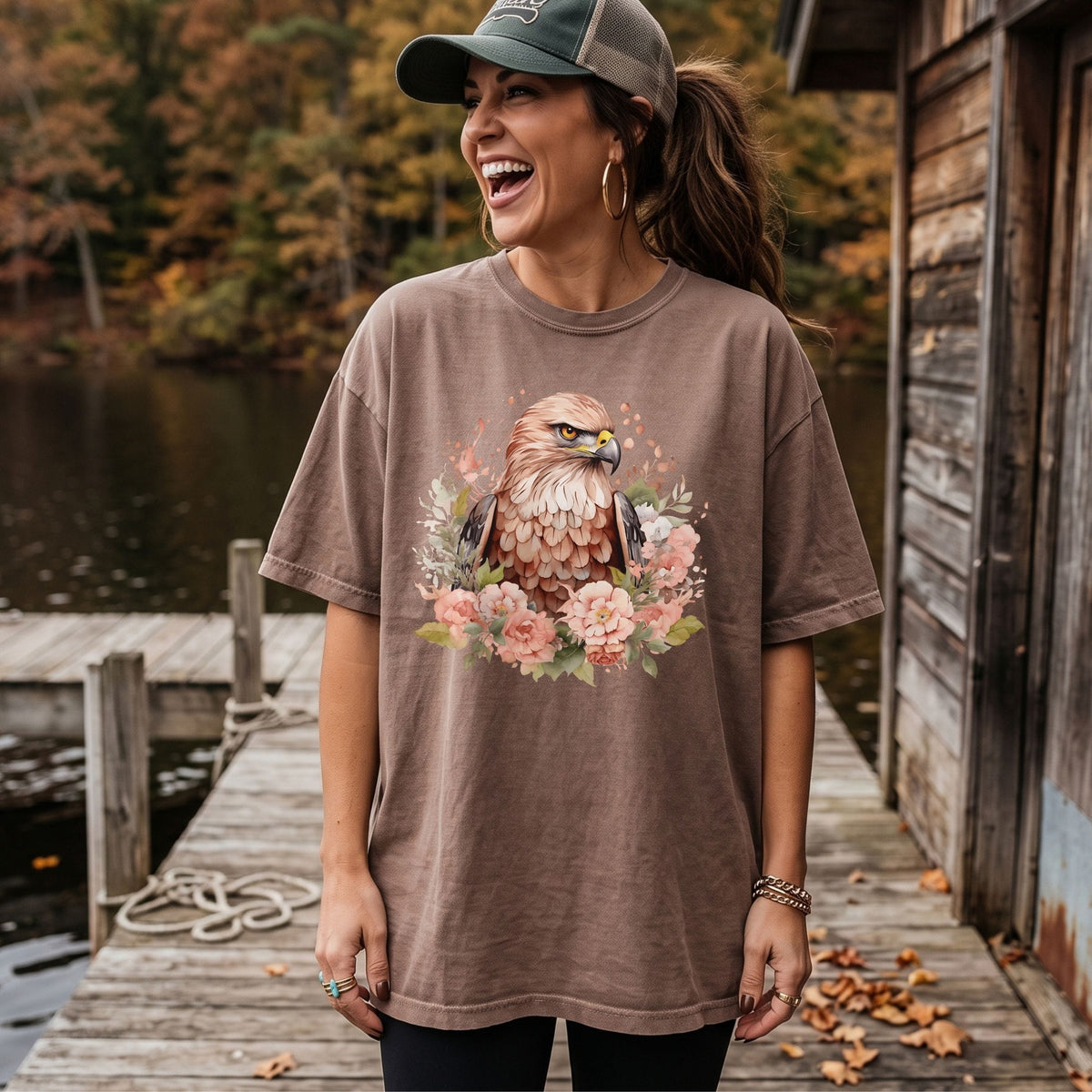A woman wearing an oversized brown t-shirt with a detailed graphic of a hawk surrounded by pink flowers, standing on a wooden dock next to a lake.