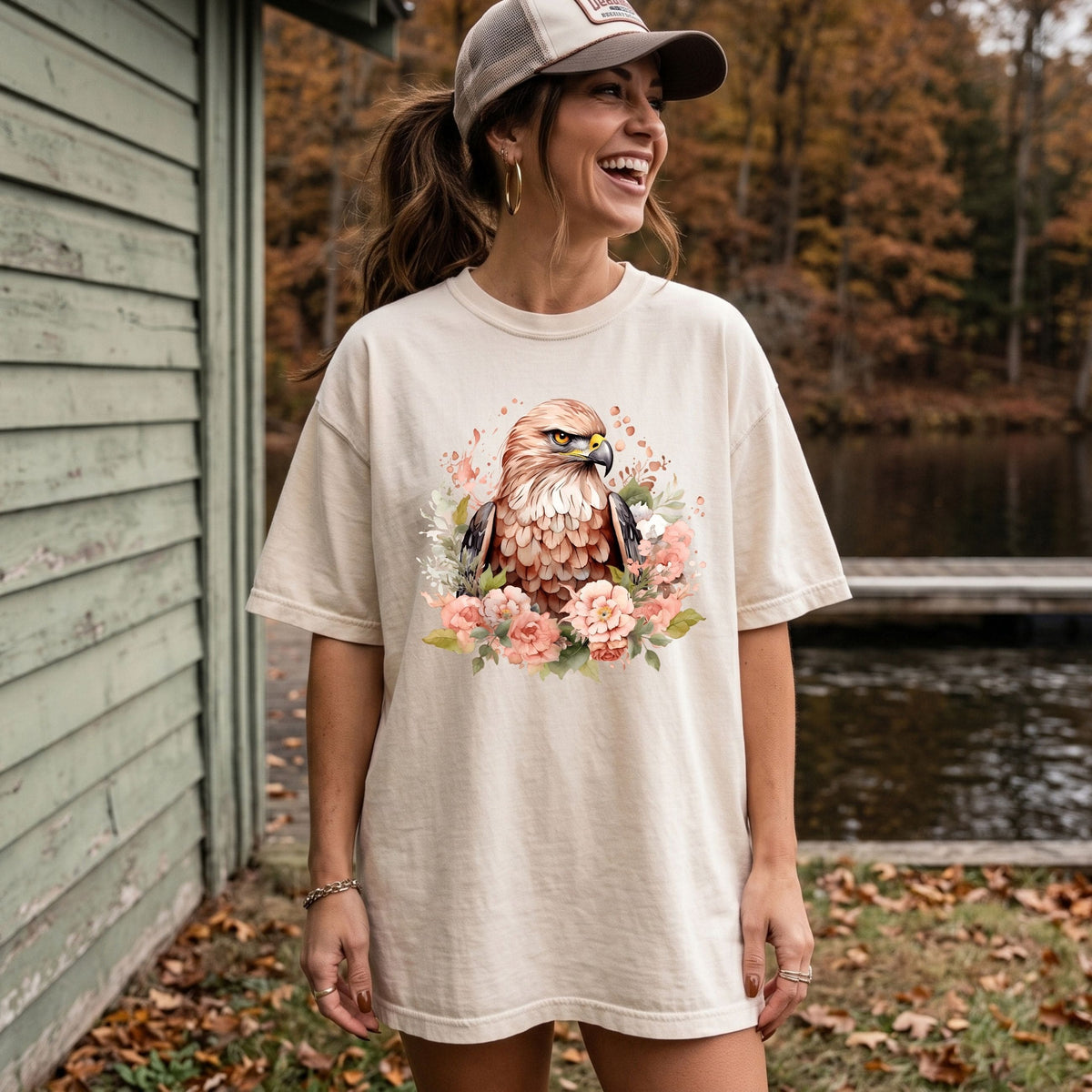 A woman wearing an oversized cream-colored t-shirt with a detailed graphic of a hawk surrounded by pink flowers, standing outdoors next to a green wooden building and a lake.