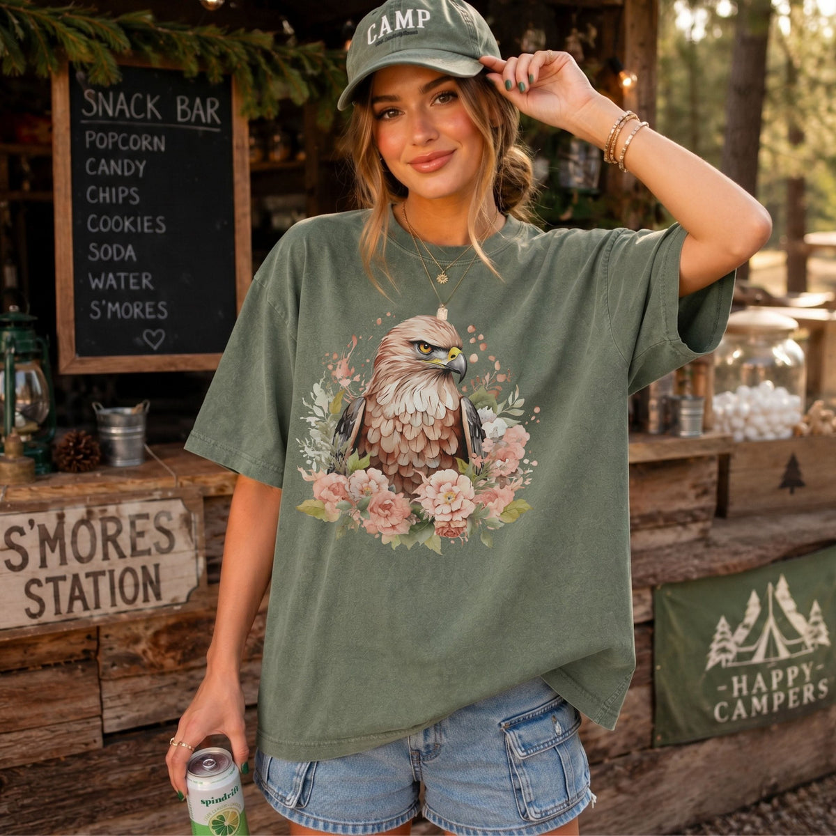 A woman wearing an oversized moss green t-shirt with a detailed graphic of a hawk surrounded by pink flowers, standing in front of a rustic wooden 'Happy Campers' snack bar.