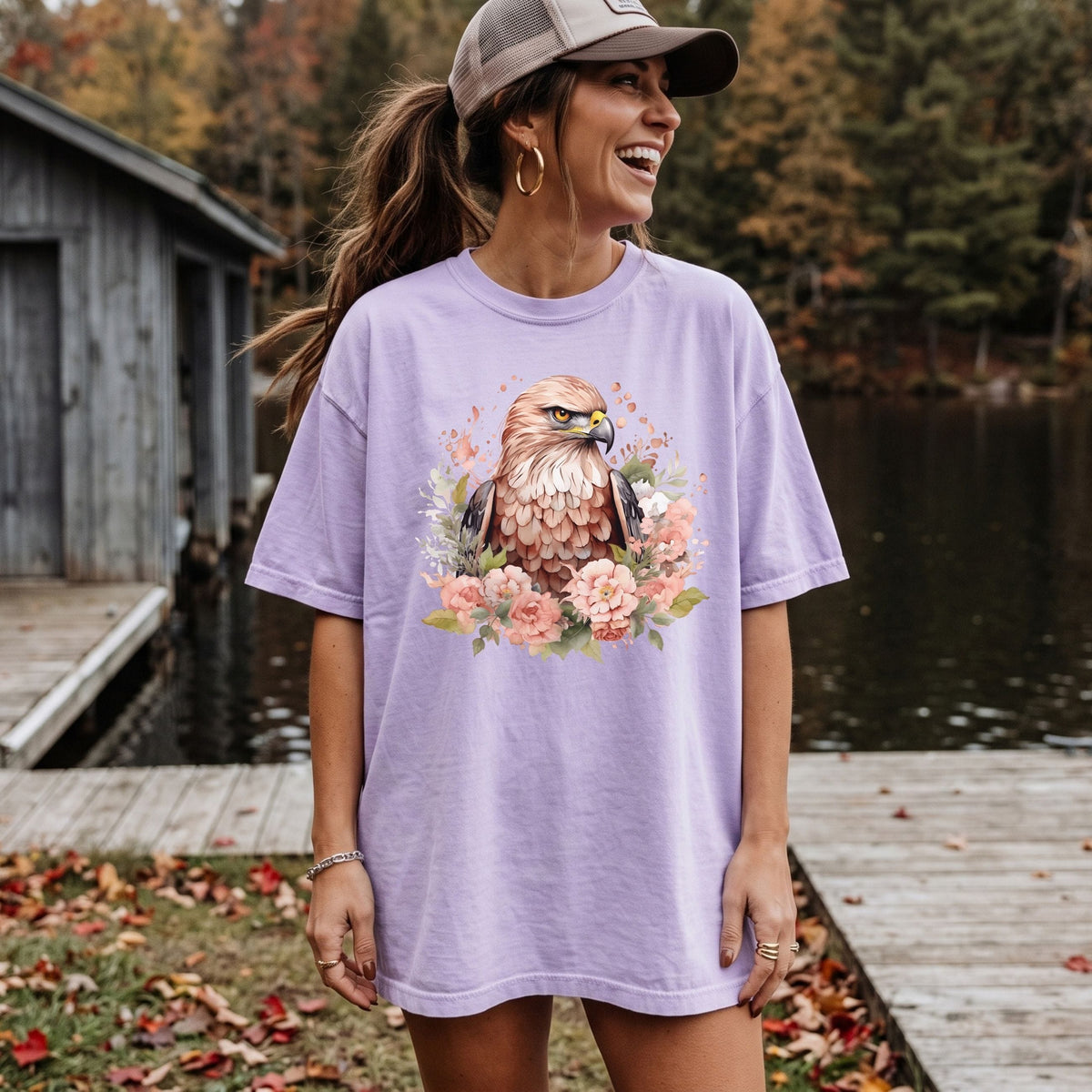 A woman wearing an oversized lavender t-shirt with a detailed graphic of a golden eagle surrounded by pink flowers, standing on a wooden dock by a lake with a cabin in the background.