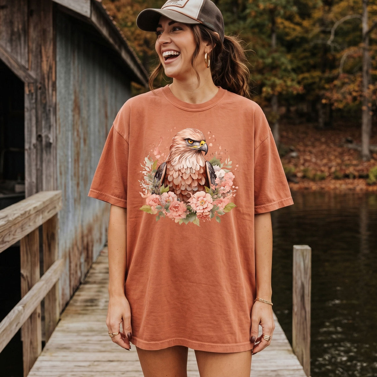 A woman wearing an oversized rust-orange t-shirt with a detailed graphic of a bald eagle surrounded by pink flowers, standing on a wooden dock next to a lake with a rustic cabin in the background.