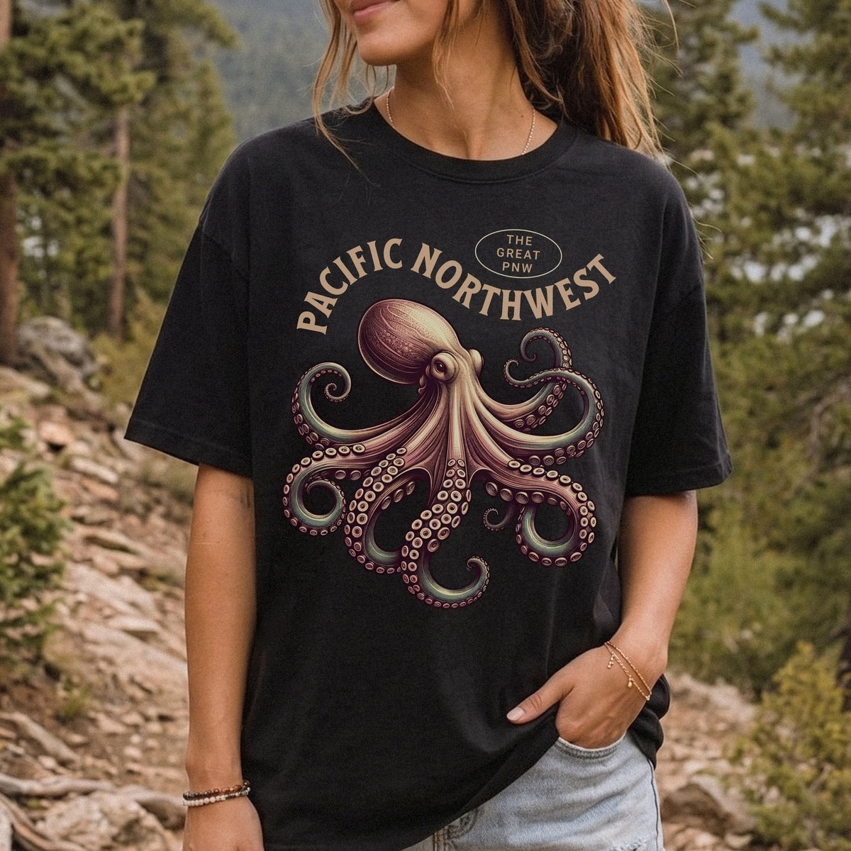 A woman wearing a black graphic t-shirt with a vintage-style octopus design and the text 'PACIFIC NORTHWEST' and 'THE GREAT PNW'. The background is a blurred outdoor forest scene.