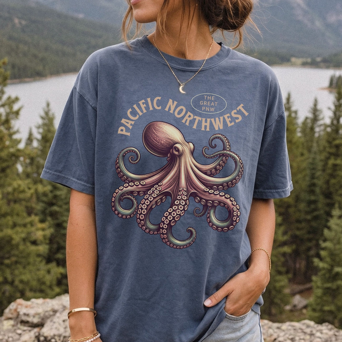 A woman wearing a faded blue graphic t-shirt with a large octopus design and the text 'PACIFIC NORTHWEST' and 'THE GREAT PNW', photographed outdoors with a mountain and lake background.