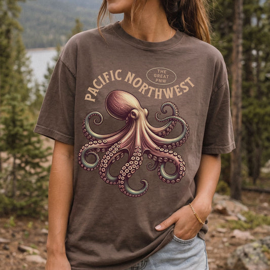 A woman wearing a brown graphic t-shirt with a vintage-style octopus illustration and the text 'PACIFIC NORTHWEST' and 'THE GREAT PNW' on the front, set against a blurred forest background.