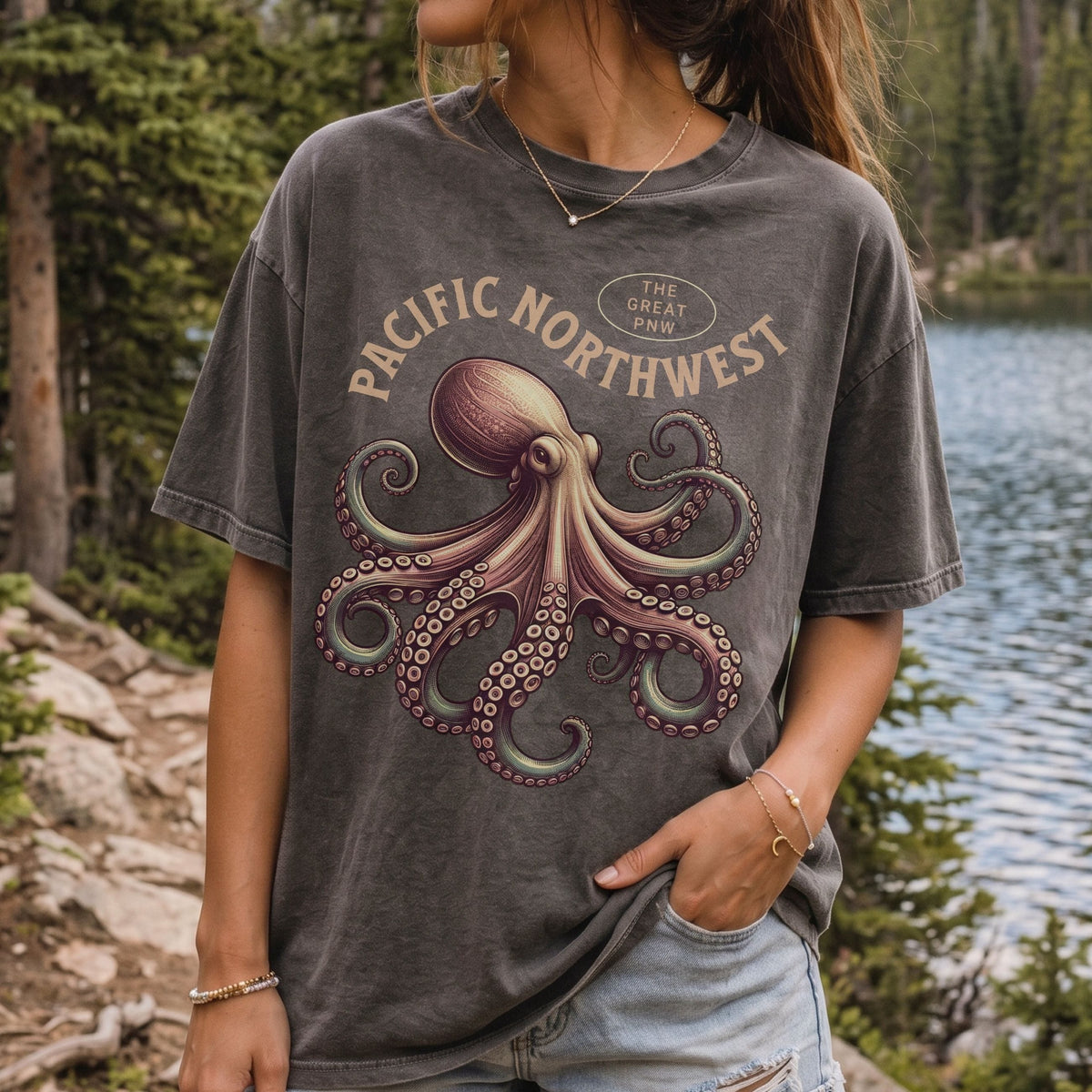 A woman wearing a faded dark gray graphic t-shirt with a vintage-style octopus design and the text 'PACIFIC NORTHWEST' and 'THE GREAT PNW'. The background shows a scenic outdoor setting with a lake and pine trees.