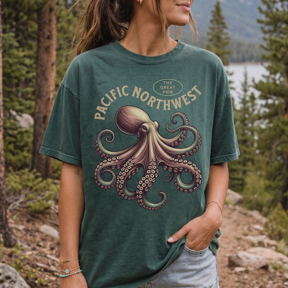 A woman wearing a dark green graphic t-shirt with a vintage-style octopus and 'Pacific Northwest' print, set against a natural forest background.