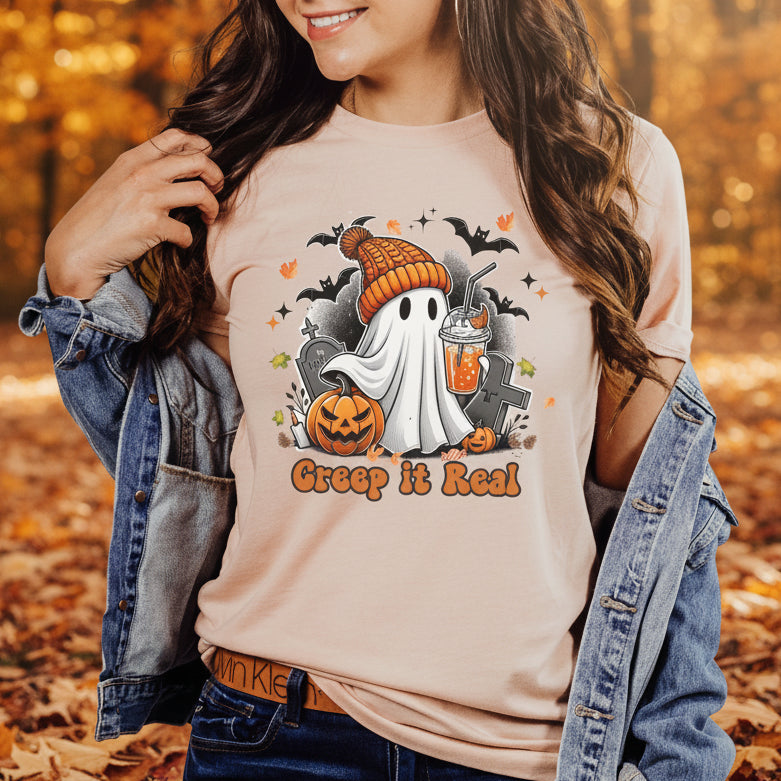 A woman wearing a light peach t-shirt with a Halloween graphic that reads 'Grep it Real' featuring a ghost, pumpkin, and tombstone, styled with an open denim jacket against an autumn leaf background.