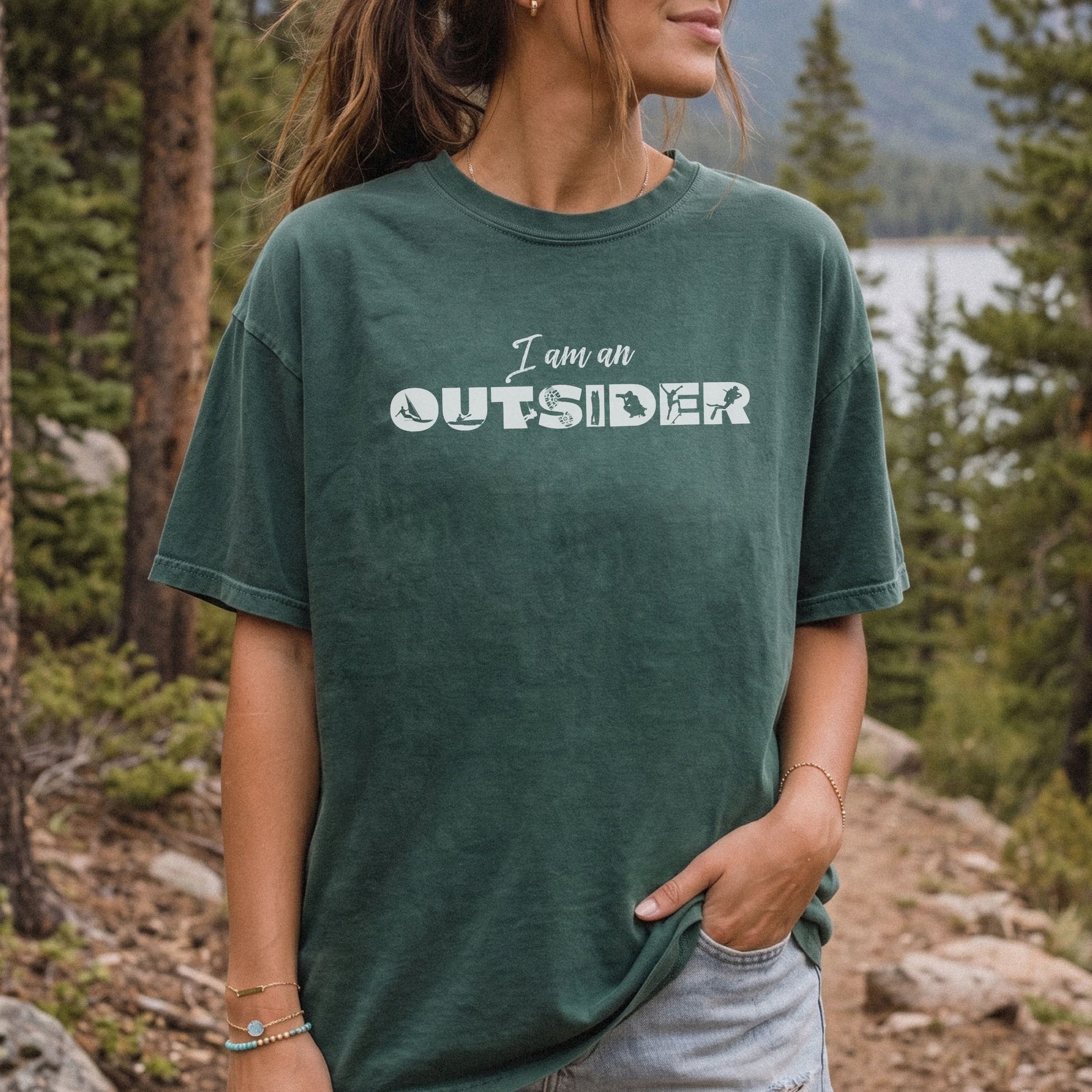 A woman wearing a dark green oversized t-shirt with the text 'I am an OUTSIDER' printed in white across the chest, set against a blurred forest and lake background.