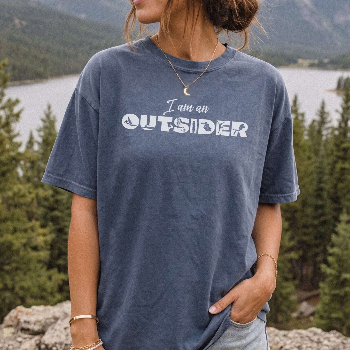 A woman wearing a faded denim blue t-shirt with the text 'I am an OUTSIDER' printed in white across the chest, set against a scenic background of a lake and pine trees.