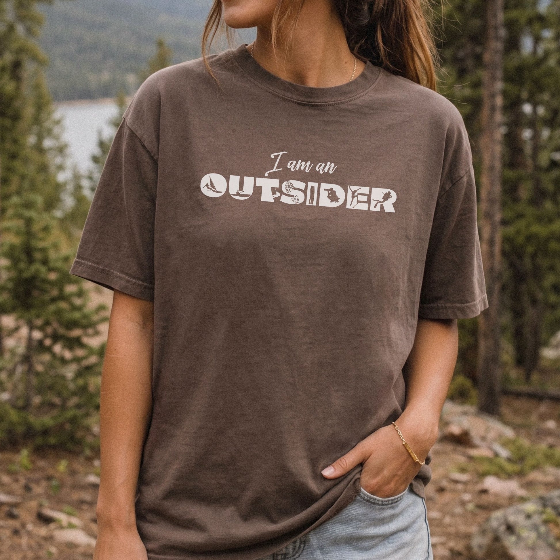 A woman wearing a brown washed graphic t-shirt with the text 'I am an OUTSIDER' printed in white across the chest, set against a blurred outdoor forest background.