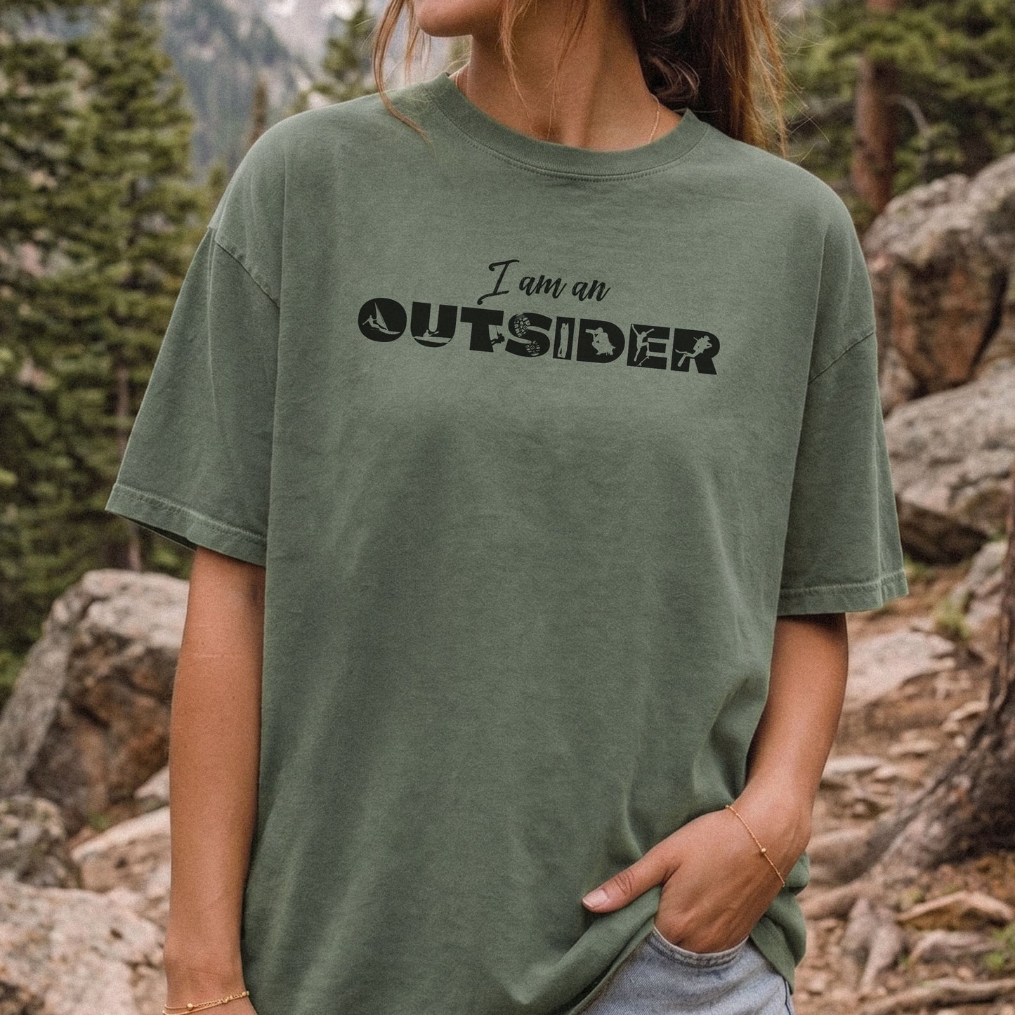 A woman wearing an oversized, faded olive green t-shirt with the text 'I am an OUTSIDER' printed in black across the chest, set against a natural outdoor mountain and forest background.