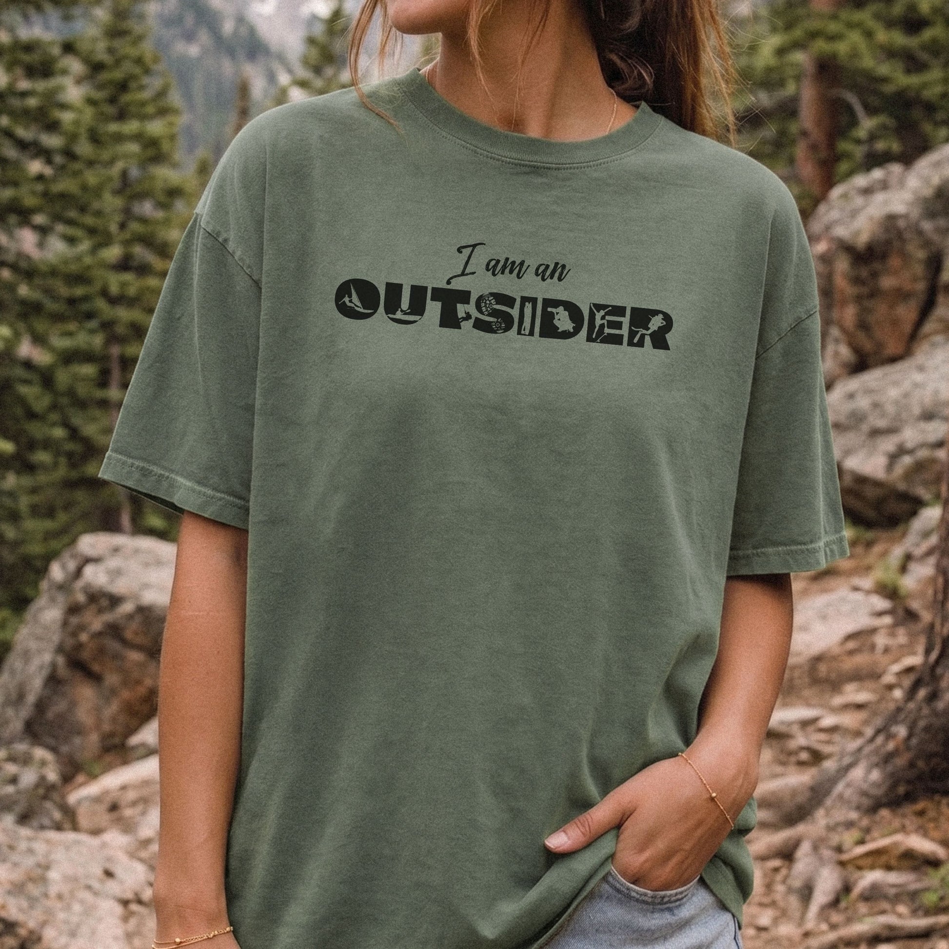 A woman wearing an oversized, faded olive green t-shirt with the text 'I am an OUTSIDER' printed in black across the chest, set against a natural outdoor mountain and forest background.