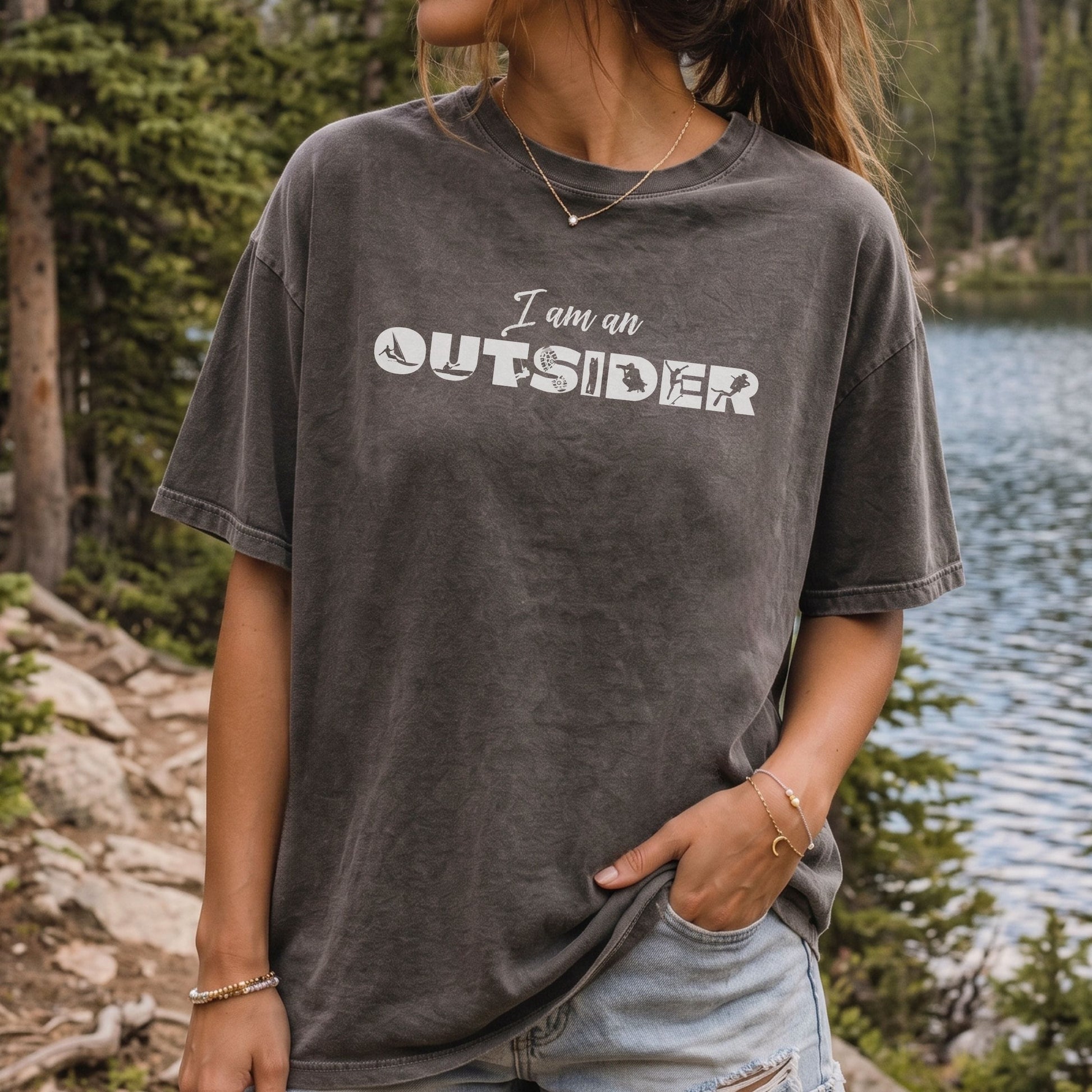 A woman wearing an oversized dark gray mineral-wash graphic t-shirt with the text 'I am an OUTSIDER' printed in white across the chest, standing outdoors near a lake and pine trees.
