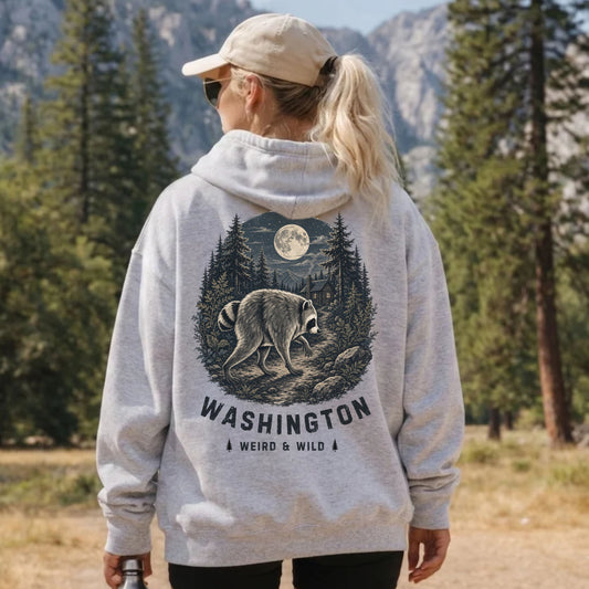 Woman wearing a light gray hoodie with a large back graphic featuring Jimothy the raccoon walking through a moonlit evergreen forest with mountains and a cabin. The design includes the words “WASHINGTON” and “WEIRD & WILD,” with a mountain landscape in the background.
