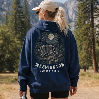 A woman wearing a navy blue hoodie with a large back graphic of a raccoon under a full moon and the text 'WASHINGTON WEIRD & WILD', photographed outdoors in a forest setting.