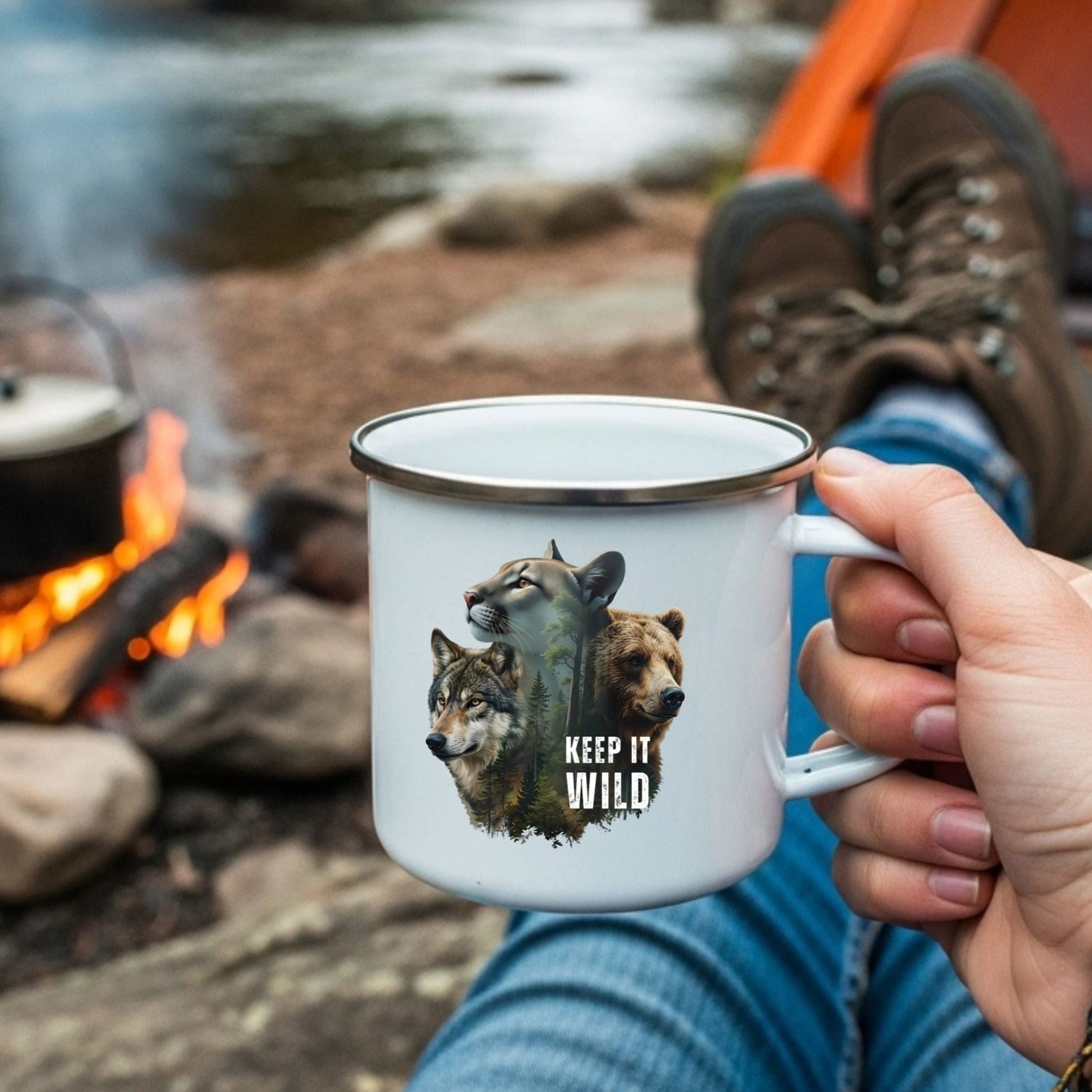 A white enamel camping mug with a silver rim is held by a person at a campsite by a river. The mug features a graphic of a mountain lion and two wolves with the text 'KEEP IT WILD'.