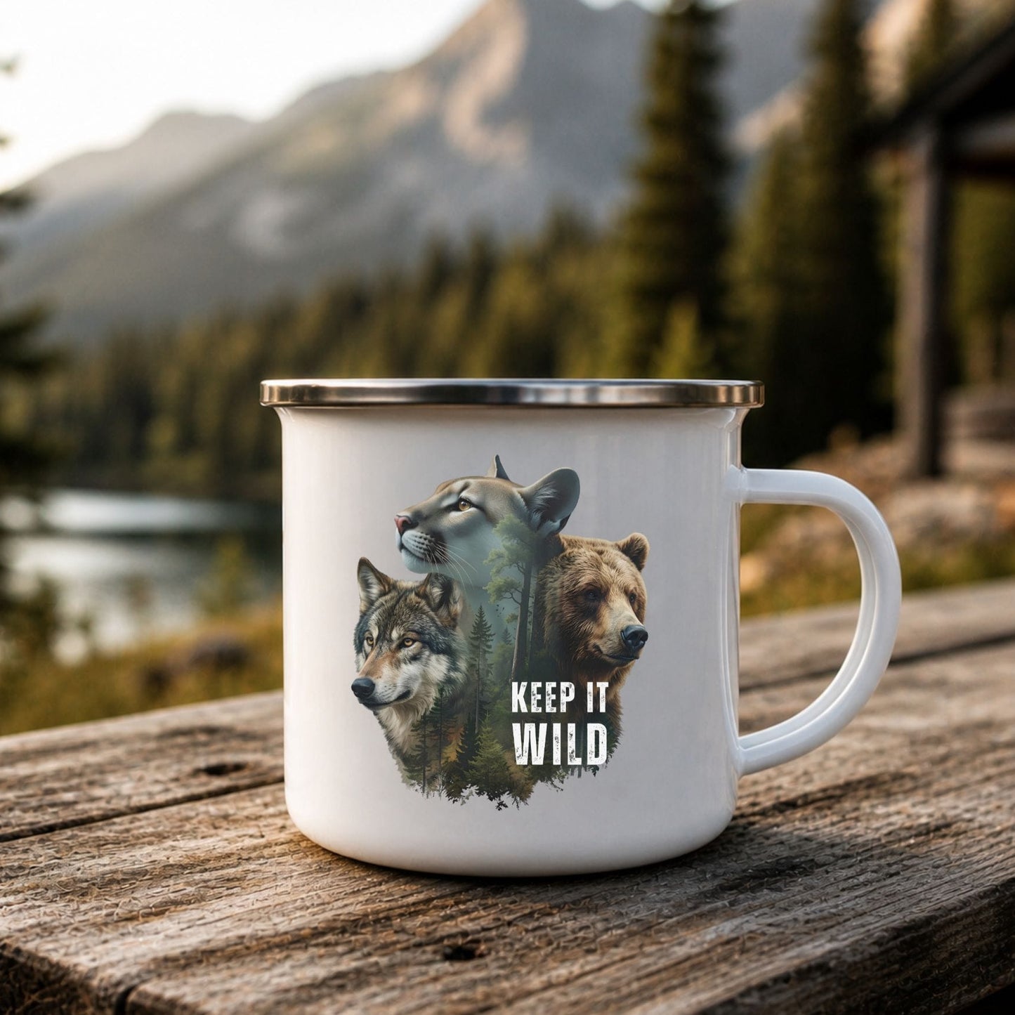 A white enamel-style mug with a silver rim sits on a rustic wooden table outdoors, with a mountain lake and pine forest in the background. The mug features a graphic of a wolf, a bear, and a cougar with the text 'KEEP IT WILD'.