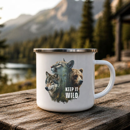 A white enamel-style mug with a silver rim sits on a rustic wooden table outdoors, with a mountain lake and pine forest in the background. The mug features a graphic of a wolf, a bear, and a cougar with the text 'KEEP IT WILD'.