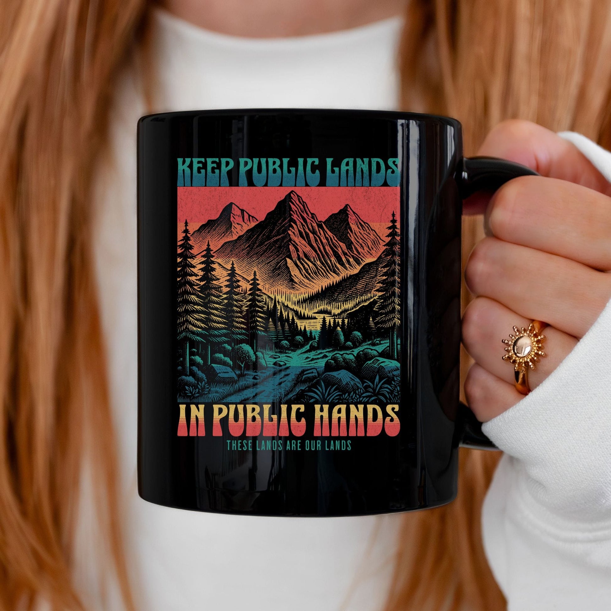 A person holding a black ceramic mug with a colorful graphic of mountains, trees, and a river, with the text 'KEEP PUBLIC LANDS IN PUBLIC HANDS'.