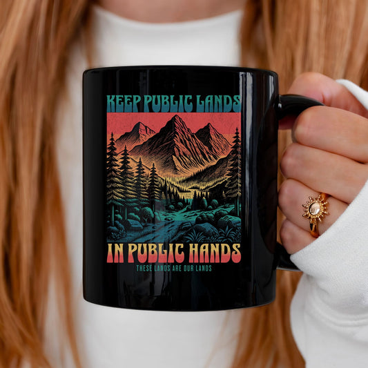 A person holding a black ceramic mug with a colorful graphic of mountains, trees, and a river, with the text 'KEEP PUBLIC LANDS IN PUBLIC HANDS'.